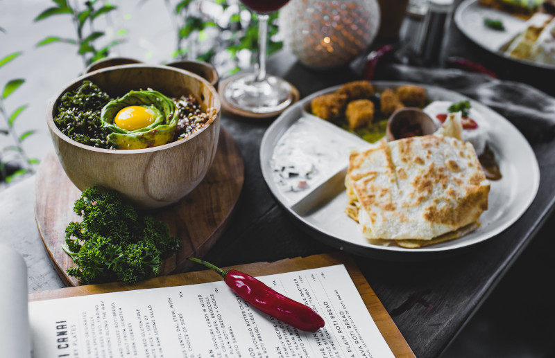 A table crowded with a bowl and a plate of fresh food, a stemmed wine glass in the background and a restaurant menu in the foreground.