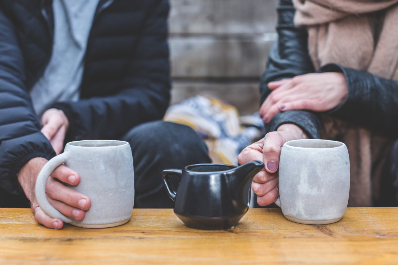 An older couple wearing coats and scarves holding grey mugs on a table, a black ceramic teapot between them.