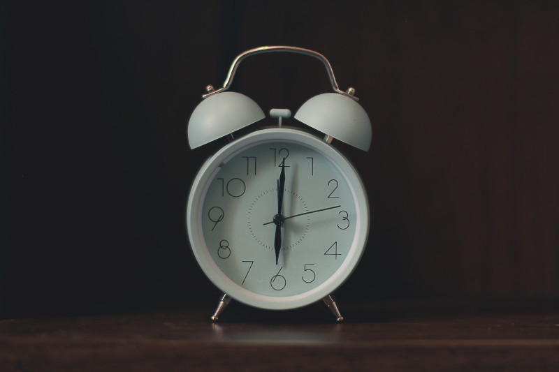 A white, analog alarm clock with two large bells on top sitting on a wooden table.