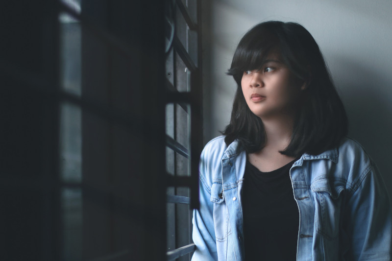 A young woman wearing a denim jacket looks through the window blinds in a shadowy room.